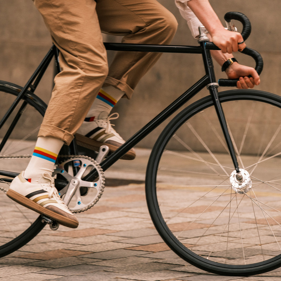 A person wearing tan pants, rainbow-striped socks, and Adidas sneakers is riding a black bicycle on a city street. The image focuses on the lower half of their body and the bikes front wheel and handlebars.