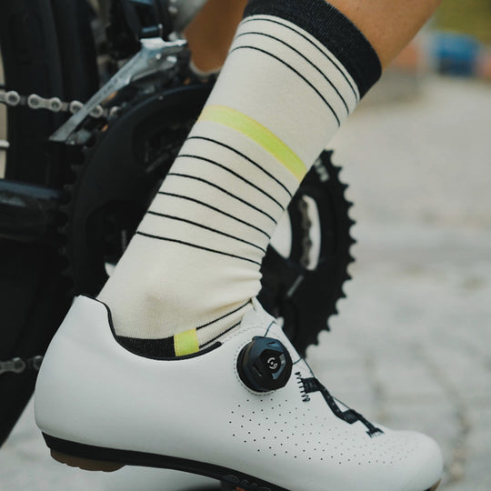 A close-up of a person’s foot wearing a white cycling shoe with a black dial and cream-colored socks with black and yellow stripes, next to a bicycle’s crankset and chainring.