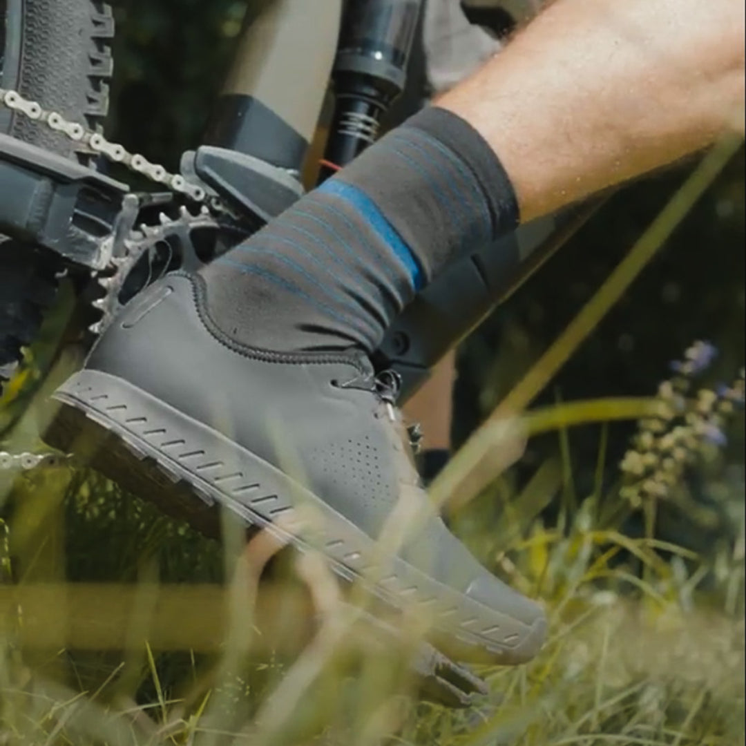 A close-up of a person’s foot on a bicycle pedal, wearing a black cycling shoe and striped socks, surrounded by green grass and plants.
