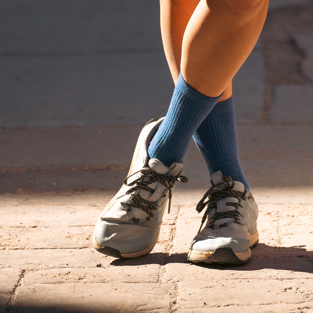 Close-up of a person standing with legs crossed, wearing blue socks and white athletic shoes with black laces on a sunlit brick surface.