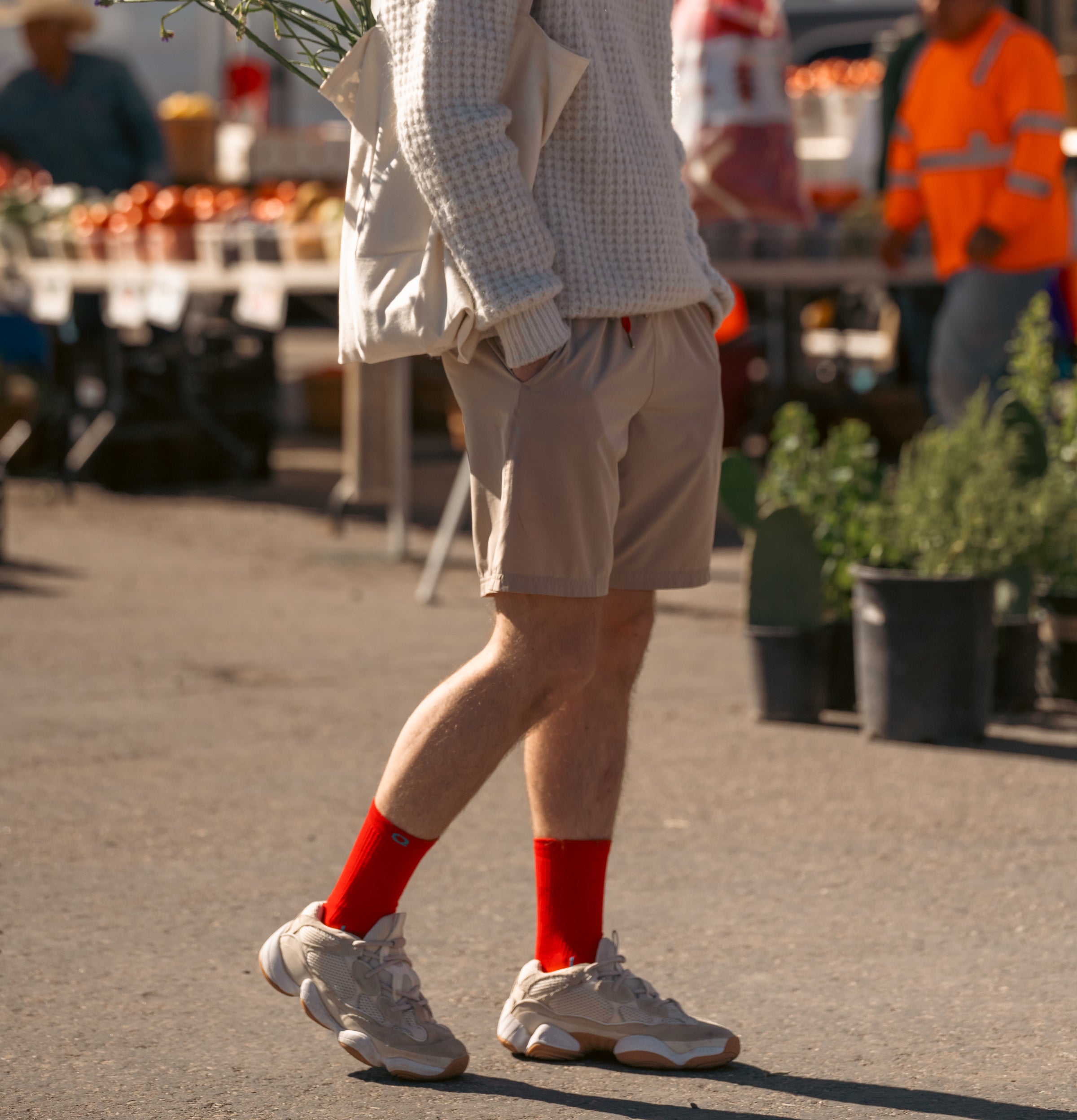 A person wearing a cream sweater, beige shorts, bright red socks, and sneakers walks at an outdoor market. The background features tables with produce and people shopping. The persons face is not visible.