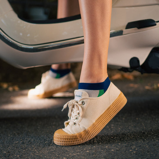 A person wearing beige canvas sneakers with thick, textured rubber soles stands on pavement beside an open car door, with part of the lower legs and shoes visible.