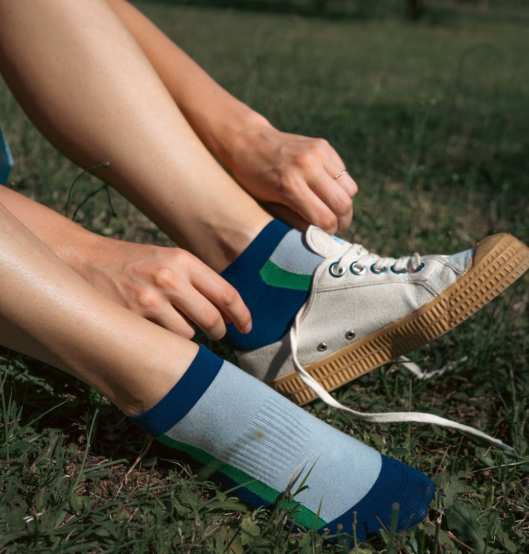 A person sitting on grass puts on a white sneaker with tan sole, already wearing a blue, white, and green sock on one foot and adjusting the other sock with both hands.