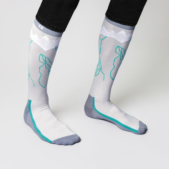 Person wearing knee-high, light gray socks with teal geometric designs and gray toes and heels. The socks feature white mountain-like patterns near the top. The person is standing on a white surface.