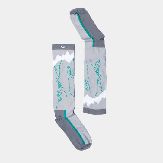 A pair of gray knee-high socks with teal and white mountain designs, laid flat on a light background. The socks feature zigzag white stripes resembling snowy peaks.
