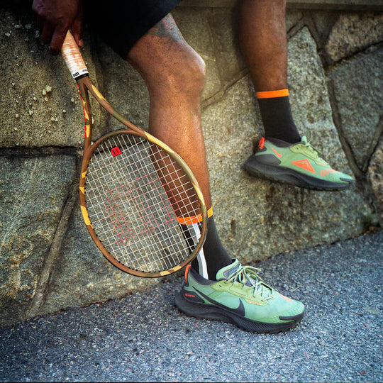 A person wearing green sneakers and black socks sits against a stone wall, holding a wooden tennis racket with one hand resting on the handle. Only their legs and part of their arm are visible.