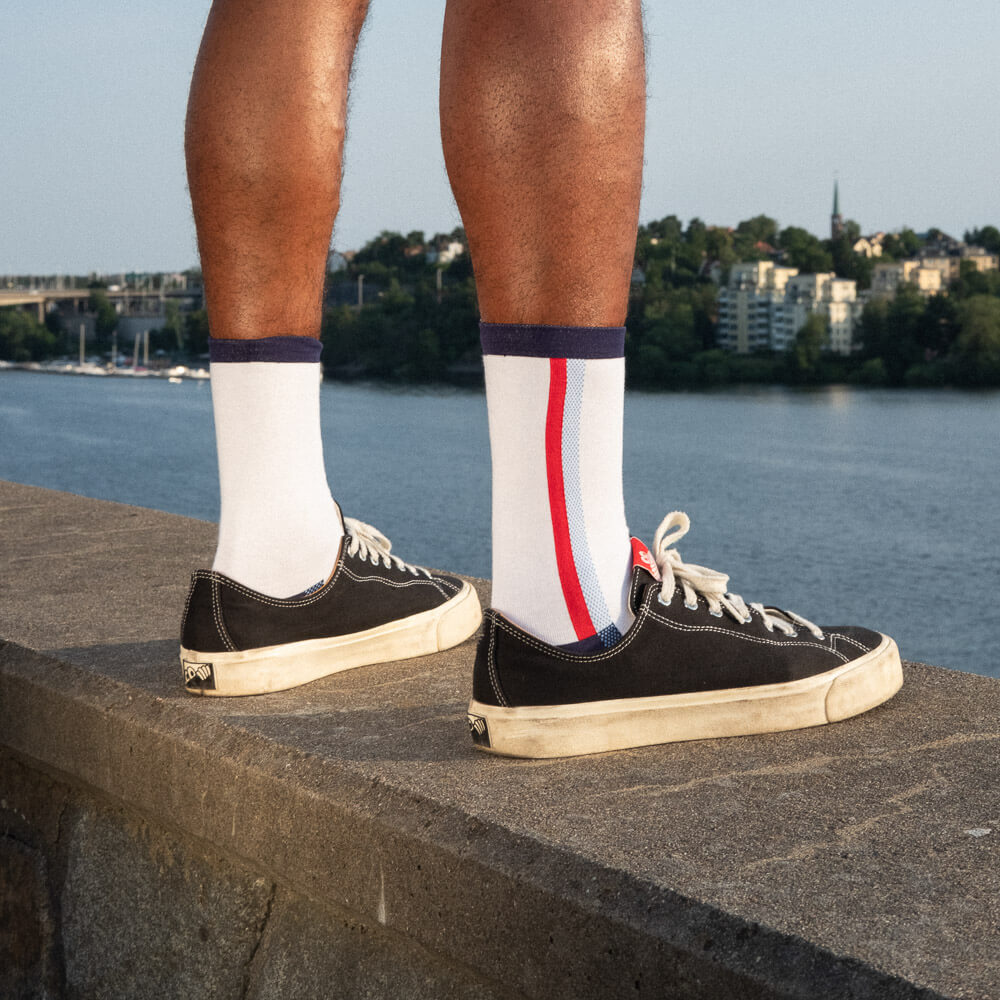 A person wearing white socks with blue and red stripes and black sneakers stands on a stone ledge overlooking a body of water, with buildings and trees visible in the background.