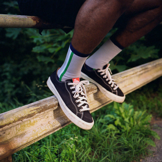 A person wearing black sneakers with white laces and striped white socks sits on a weathered wooden fence, surrounded by green foliage.