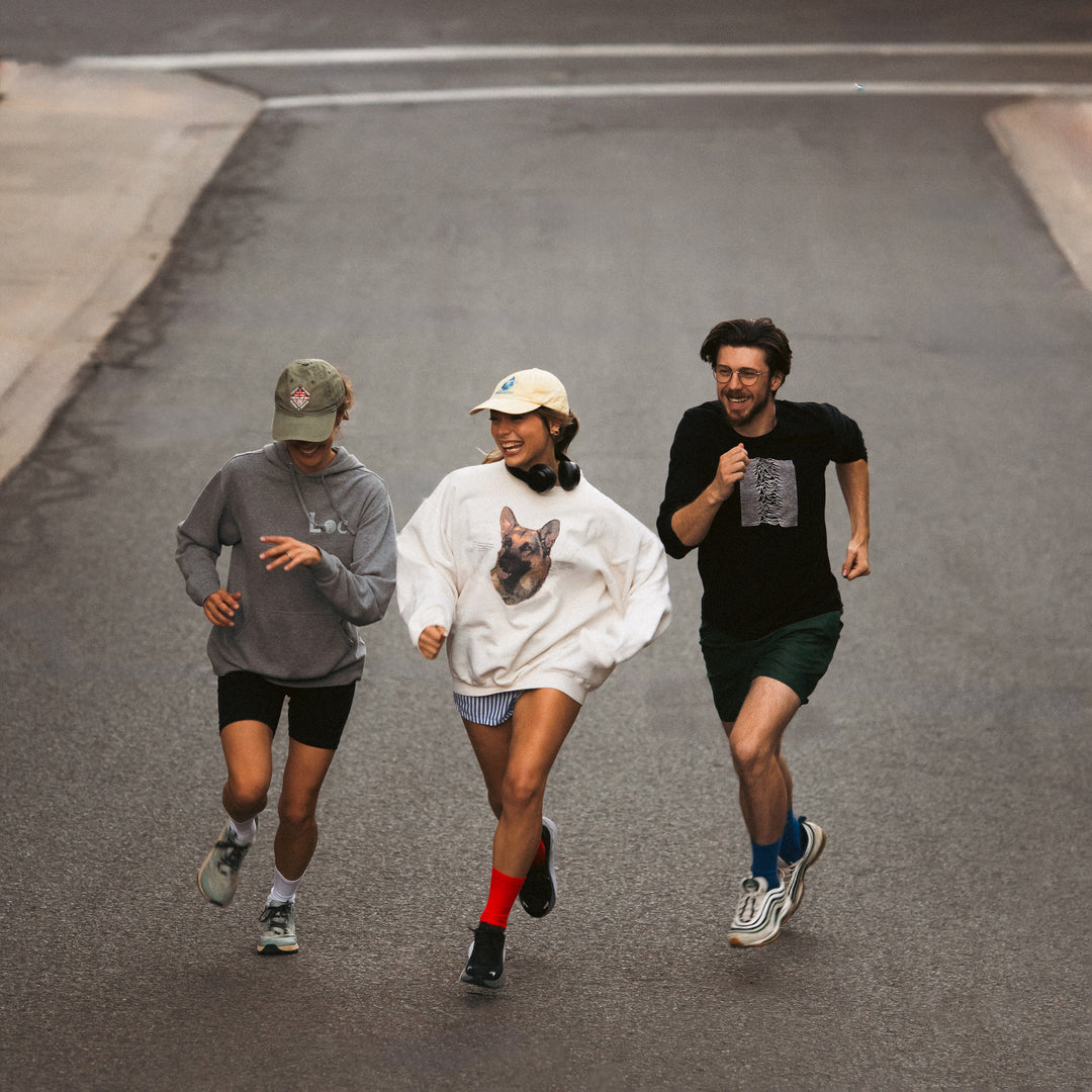Three people wearing casual athletic clothing jog together on a paved street, smiling and enjoying the activity. One person wears a white sweatshirt with a dog graphic, and all appear relaxed and happy.