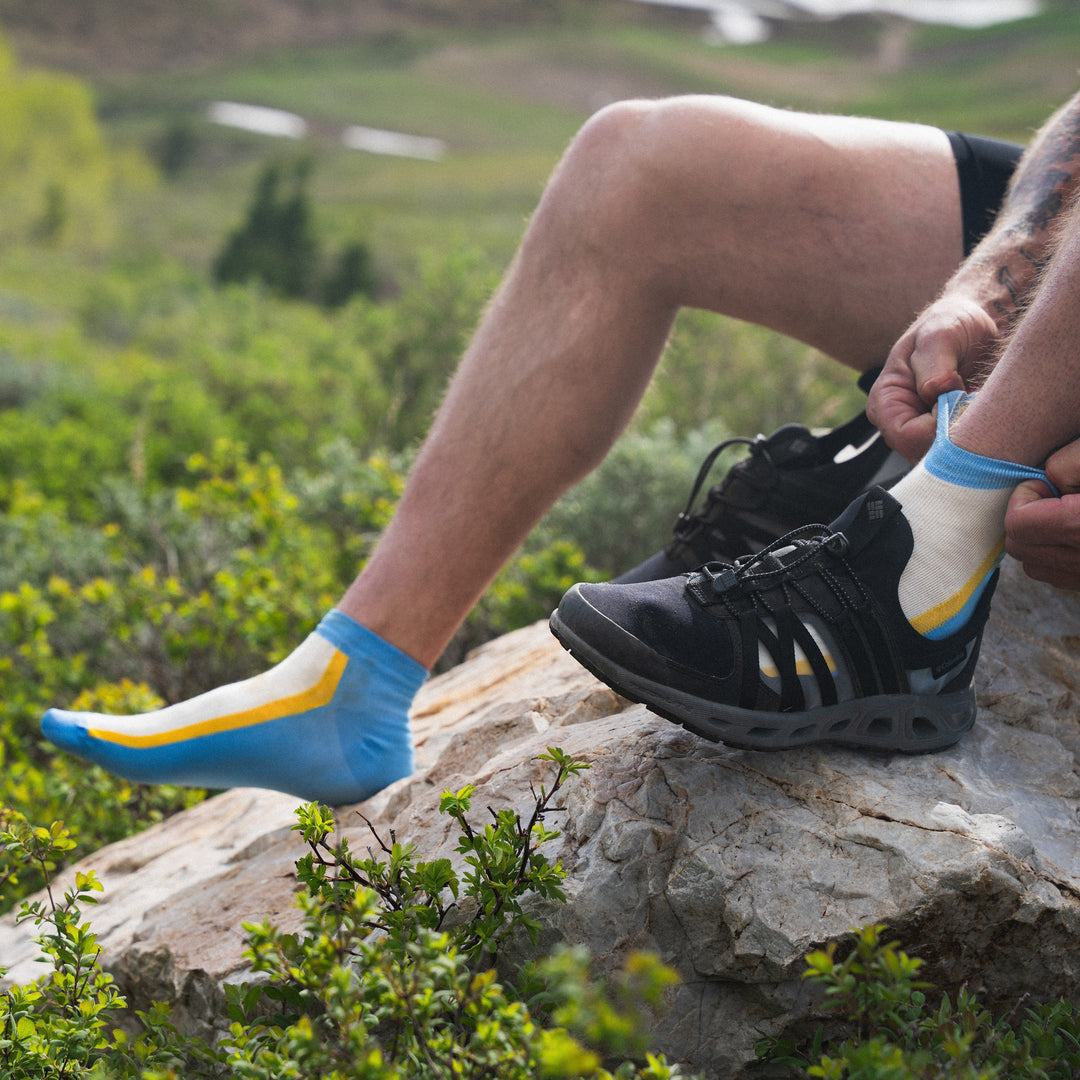 A person sits on a rock outdoors, wearing one black hiking shoe and adjusting a colorful blue, yellow, and white sock on their other foot, surrounded by green vegetation and hills in the background.