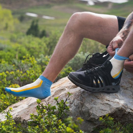 A person sits on a rock outdoors, wearing one black hiking shoe and adjusting a colorful blue, yellow, and white sock on their other foot, surrounded by green vegetation and hills in the background.