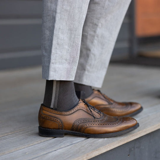 A person wearing light gray trousers, dark socks, and brown leather wingtip dress shoes stands on a wooden floor. The photo focuses on the lower legs and feet.