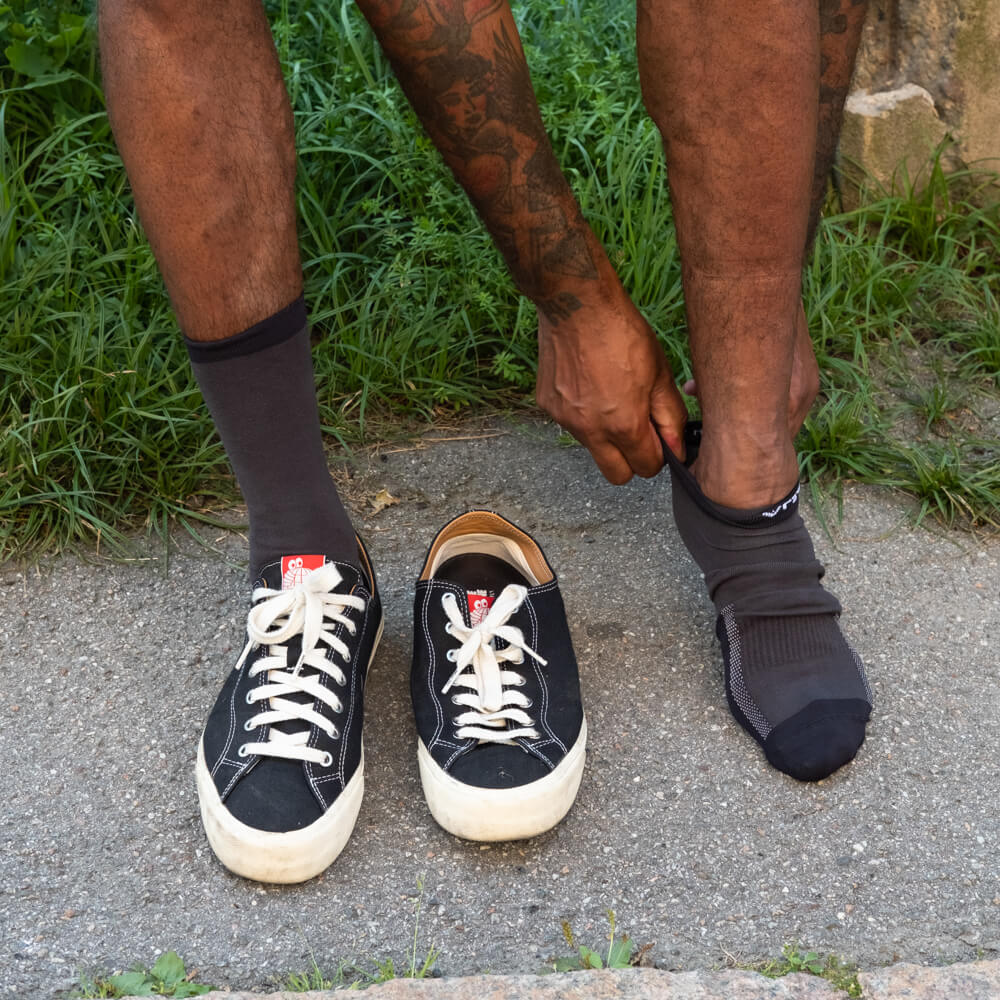 A person with tattooed arms puts on a black sock next to a pair of black sneakers with white laces, standing on a sidewalk with grass in the background.