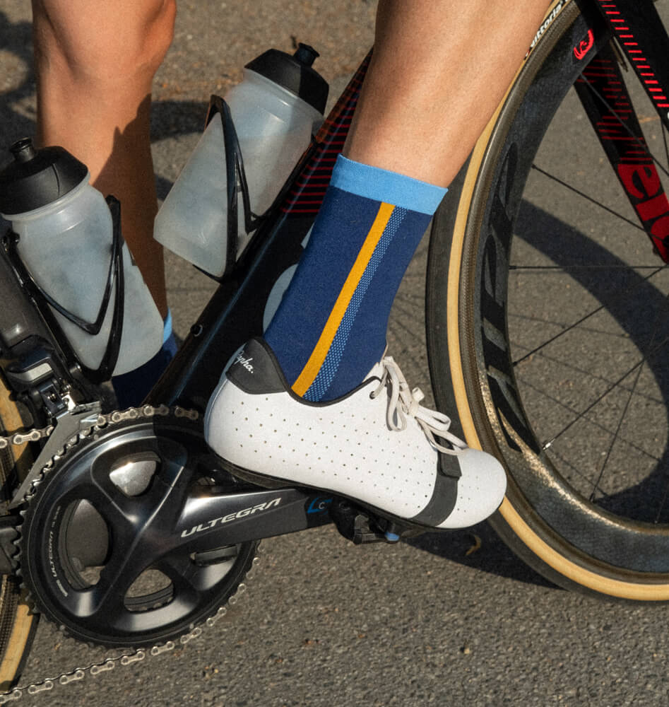 A close-up of a cyclist’s foot on a pedal, wearing a white cycling shoe and blue sock with a yellow stripe. Two water bottles are mounted on the bicycle frame, and the bike is on a paved road.