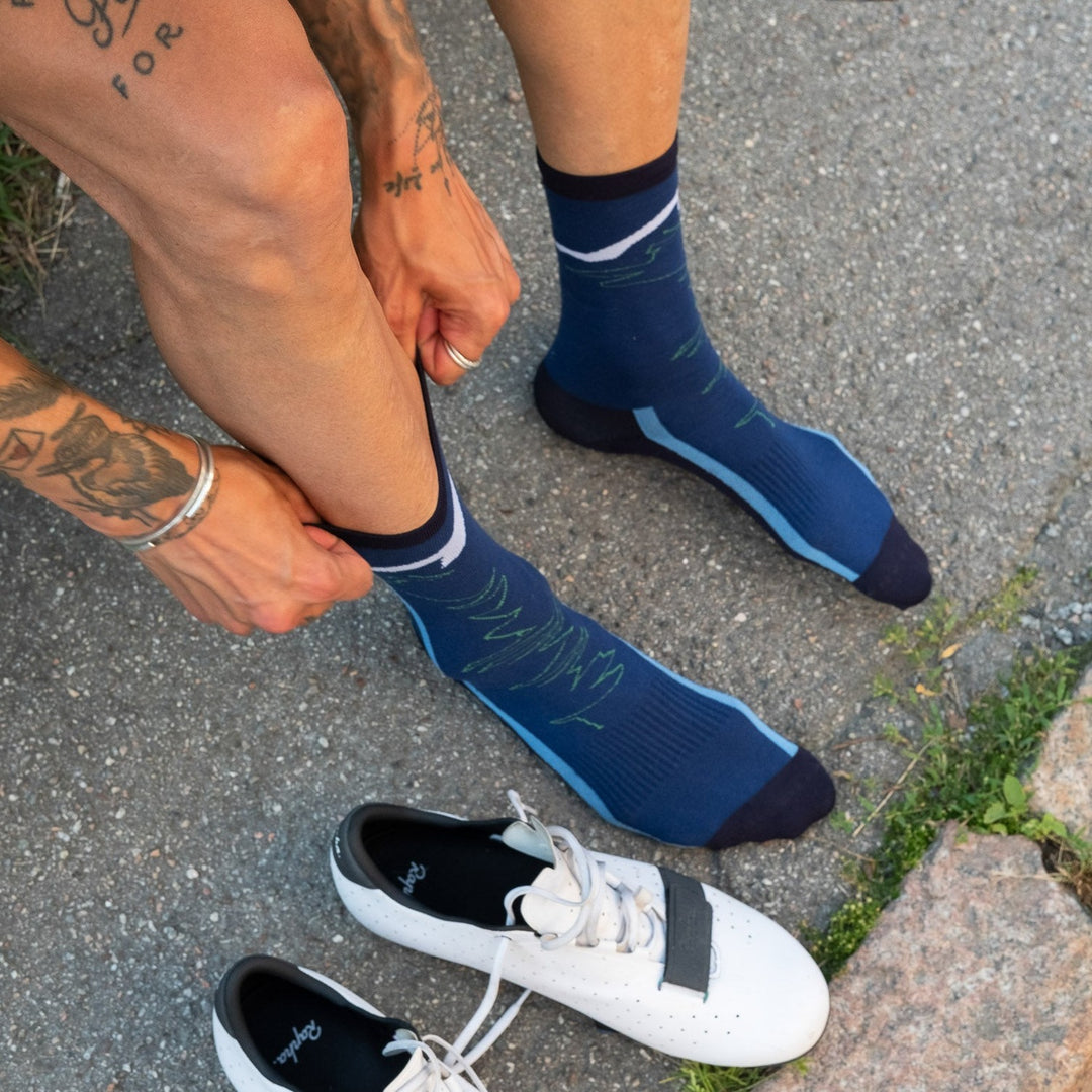 A person with tattoos on their arms and legs pulls up blue cycling socks while sitting on the ground next to a pair of white cycling shoes on a paved surface.