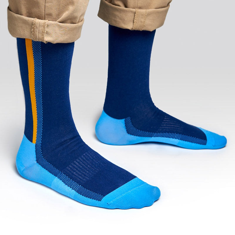 A person wearing blue and light blue socks with a yellow stripe, standing on a light gray surface. Their pants are rolled up to reveal the socks.